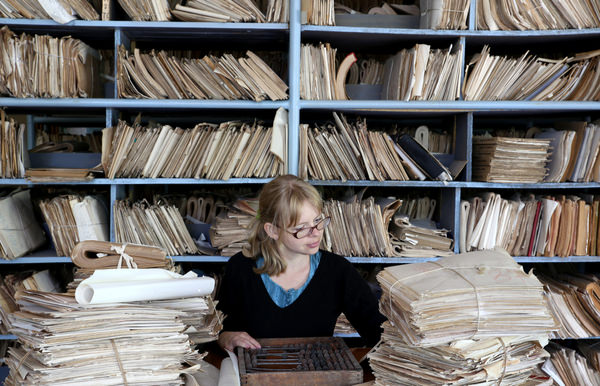 A woman works at her desk which is covered in files and folders. Shelves are filled with files behind her as well.
